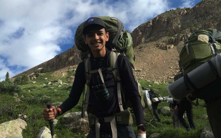 a young person wearing a backpack smiles at the camera amidst a mountainous landscape